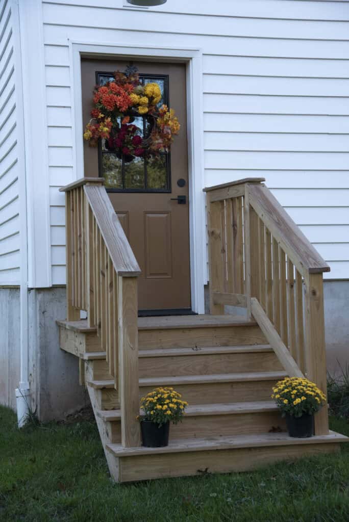 A brown door with a fall wreath is at the top of wooden steps with railings. Two pots of yellow flowers sit on each side of the bottom step. The house has white siding and green grass in front.