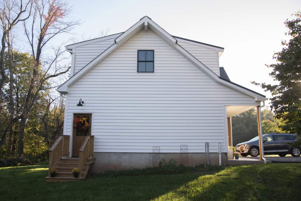 A white house with horizontal siding, a small front porch decorated with a fall wreath and potted flowers, sits on a green lawn. A car is parked under a carport to the right. Trees surround the house.