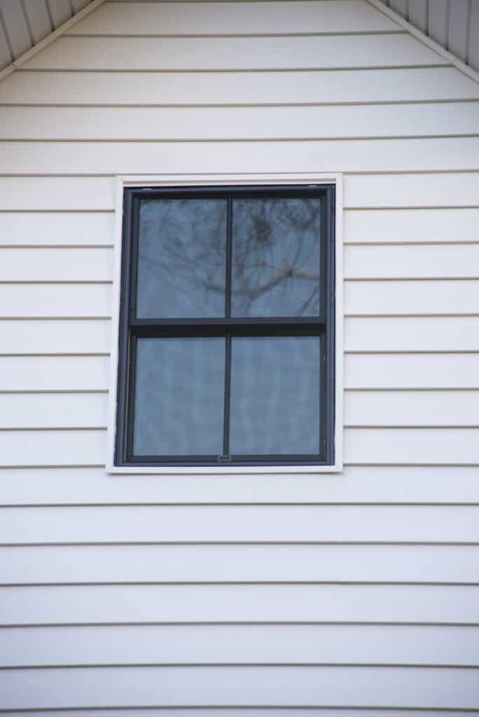 A single black-framed window with four panes is set in the white vinyl siding of a house, reflecting tree branches in the glass.