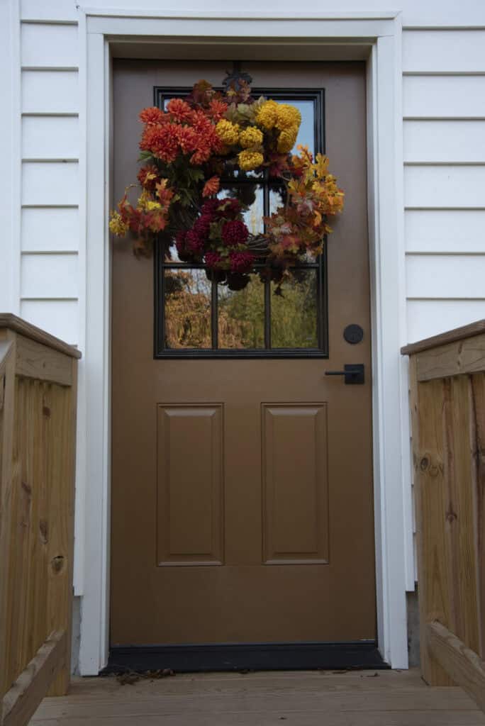 A brown front door with a window is decorated with an autumn wreath made of red, orange, and yellow flowers. The door is framed by white siding and wooden railings on both sides.