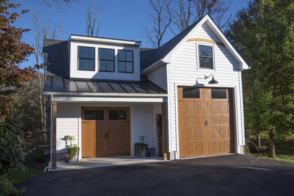 A modern two-story garage with white siding, large windows, and two wooden garage doors—one single and one double—surrounded by trees and a clear blue sky.