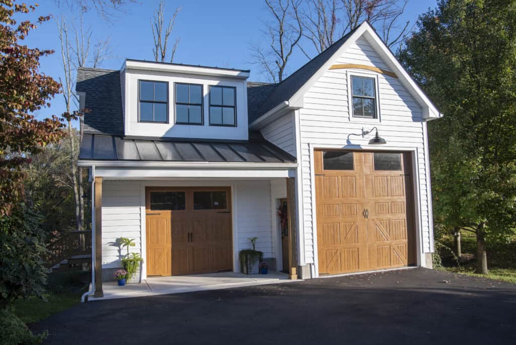 A modern white garage with two wooden doors, one large and one standard-sized, sits on a paved driveway. The building has a dormer with three windows, black roof, and is surrounded by trees under a clear blue sky.