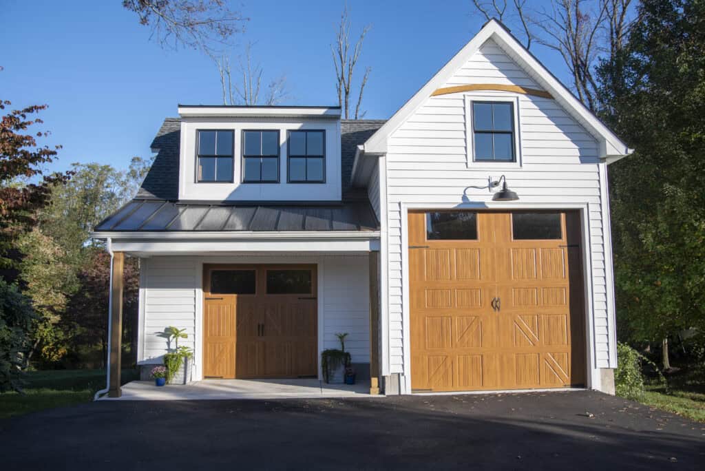 A modern two-story white garage with two wooden doors, one large and one smaller, black-framed windows, and potted plants by the entrances, set against a backdrop of trees and a clear sky.