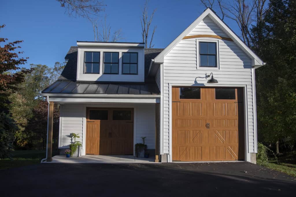 A white detached garage with two large wooden doors, one for cars and a taller one for RVs, sits on a paved driveway surrounded by trees and plants under a clear blue sky.