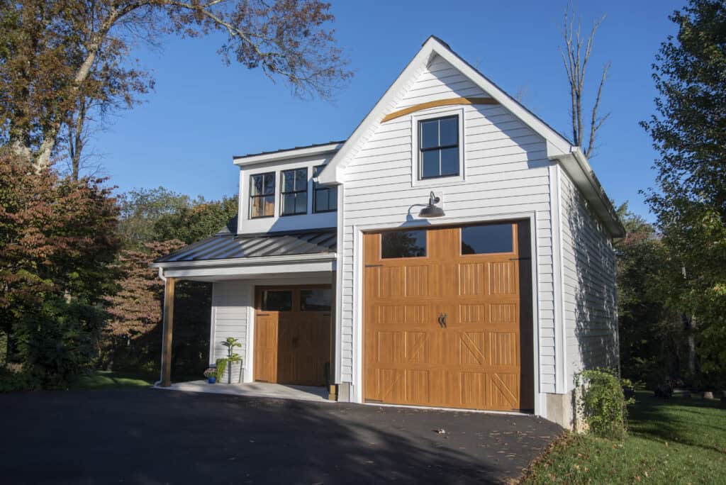A two-story white garage with wooden doors, black roof trim, and a driveway, surrounded by trees and greenery under a clear blue sky.