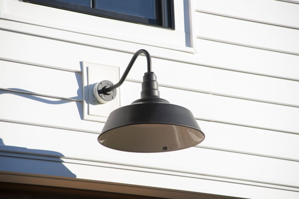 A black outdoor wall light fixture is mounted on white horizontal siding beneath a window, casting a distinct shadow on the sunlit wall.
