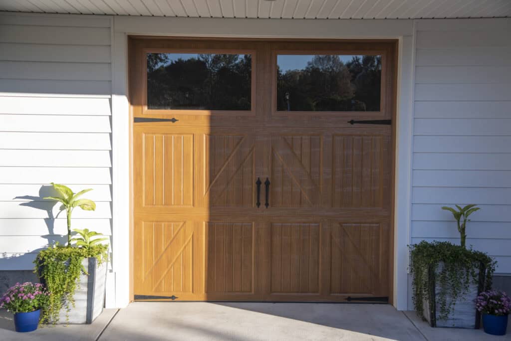 A wooden garage door with decorative black hardware, set in a white paneled exterior wall. Potted green plants and purple flowers are arranged on either side of the garage door.
