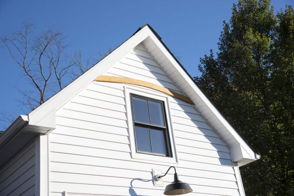 The upper part of a white house with black-trimmed window, black outdoor light fixture, and a wooden accent under the peak of the roof, against a clear blue sky and trees.