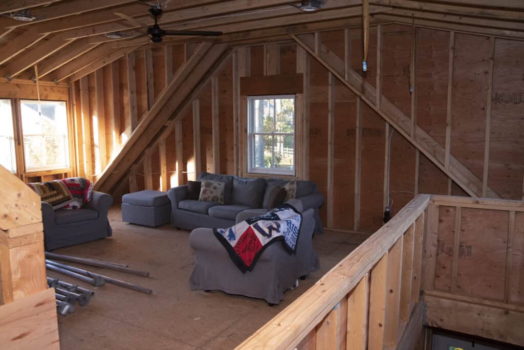 A partially finished attic with exposed wooden beams and walls, containing a gray sectional sofa with blankets and pillows, and a window letting in natural light.