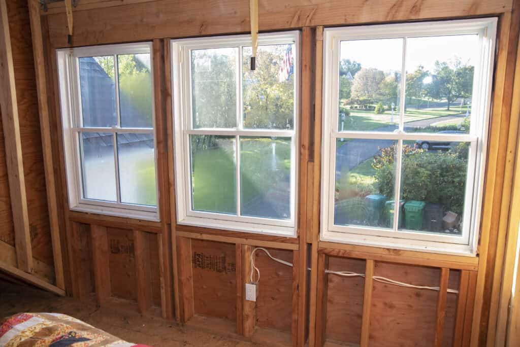 Two large windows set in an unfinished wooden wall let sunlight into a room under construction. Outside, a grassy yard, trees, street, and houses are visible. Electrical wires and outlets are exposed beneath the windows.