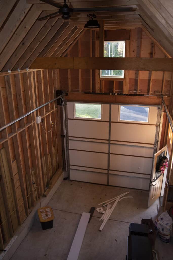 An unfinished garage with exposed wooden beams and a partially open door, letting in sunlight. Construction materials, tools, and a yellow bin are scattered on the concrete floor. A small window is above the garage door.