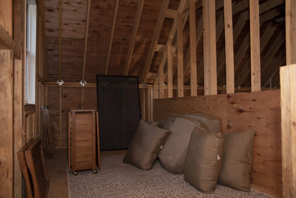 An unfinished attic with exposed wooden beams, a patterned rug, several large brown cushions, folded wooden tables, and a standing dark brown cabinet against the wall. Natural light comes in from a side window.
