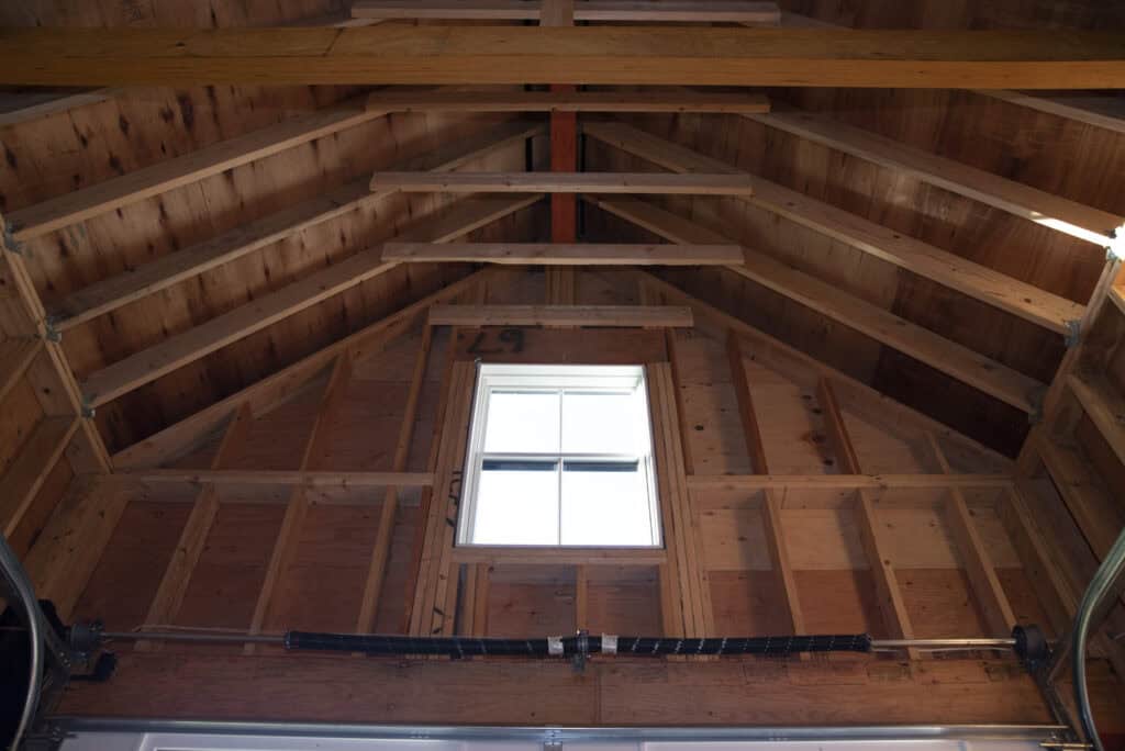 Interior view of a wooden garage or shed roof with exposed wooden beams, unfinished walls, and a small rectangular window centered on the upper wall letting in natural light.