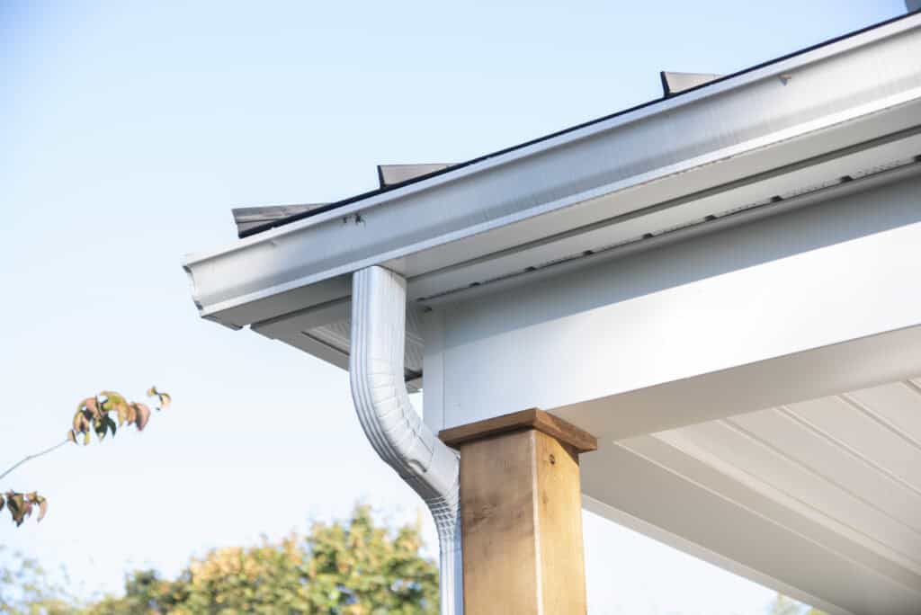 Close-up of a house roof corner with a white rain gutter and downspout attached to a wooden post, against a clear blue sky and some trees in the background.