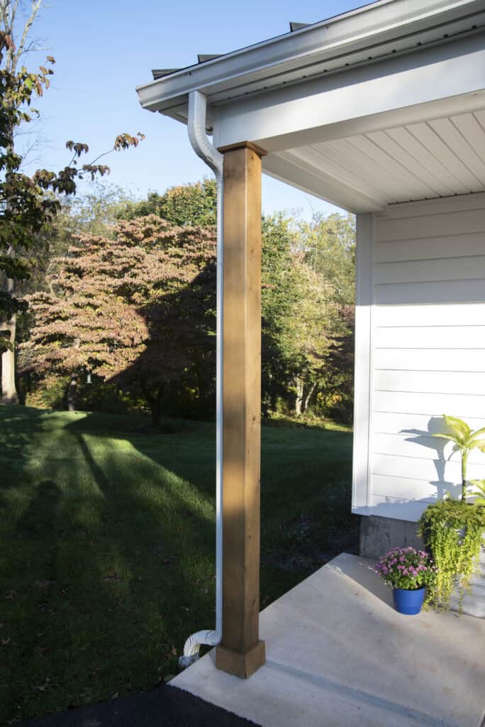 A wooden support post stands at the corner of a white house with a rain gutter above, next to a concrete porch. Green grass, trees, and a potted plant in a blue container are visible in the background.