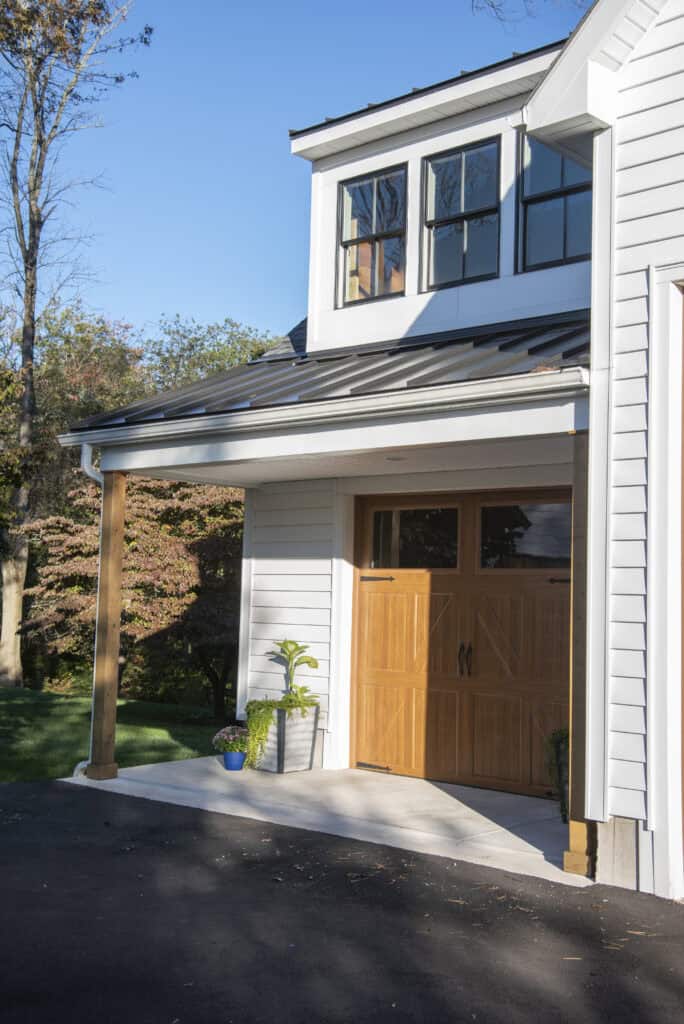 A modern white house with wooden garage doors, large windows, and a small porch with potted plants. The driveway is paved and trees are visible in the background under a clear blue sky.
