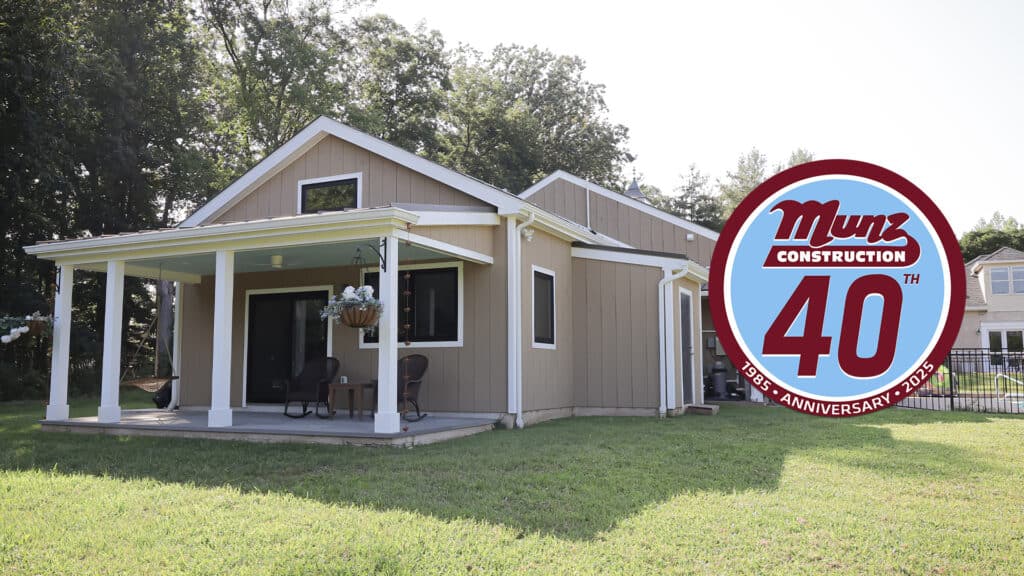A tan house with a covered porch and white trim sits on a green lawn under a sunny sky. Imagine how adding a portico could enhance its charm. A large circular logo reads Munz Construction 40th Anniversary 1985-2025 in the foreground.