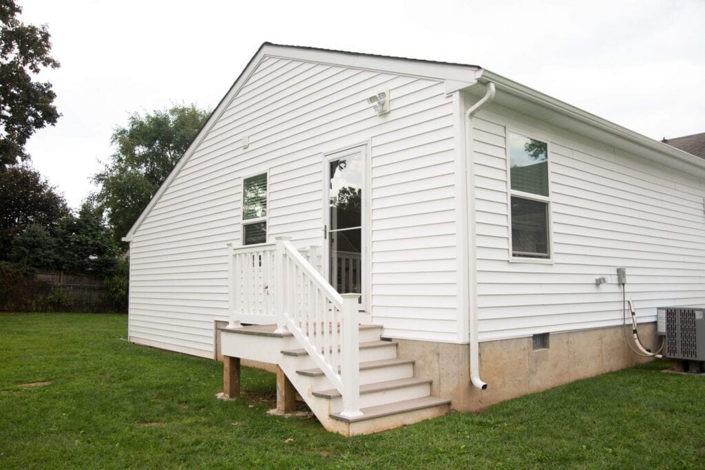 A white, single-story house with vinyl siding features a small back porch, white railing, and steps leading to a grassy yard—ideal for those exploring how to add an in-law suite or expand living space. Two windows, a door, and an outdoor AC unit are visible.