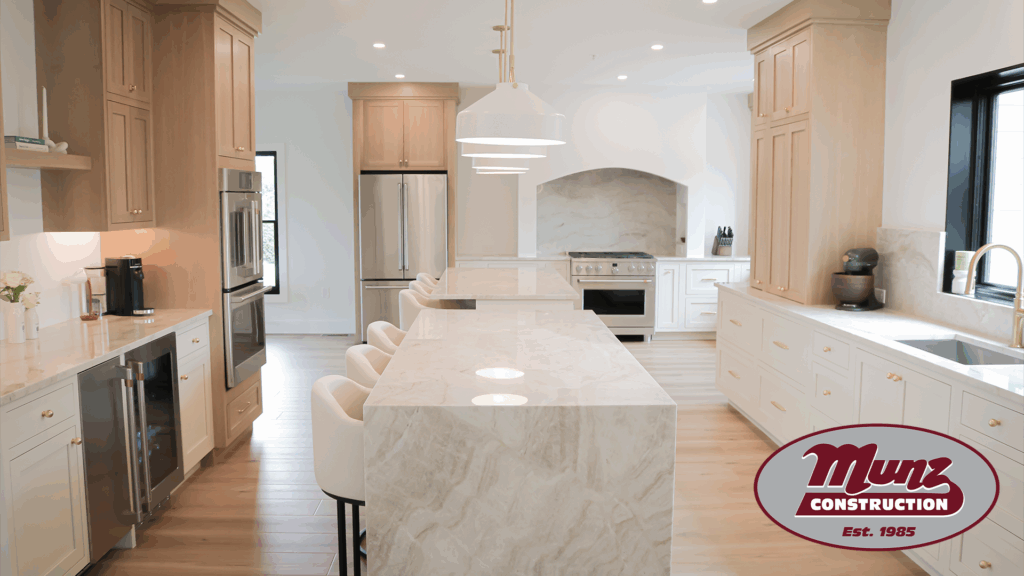 Spacious modern kitchen with marble island, white cabinets, stainless steel appliances, light wood accents, and pendant lighting—perfect inspiration if you're exploring how to build an in-law suite in Pennsylvania. Munz Construction logo appears in the lower right corner.