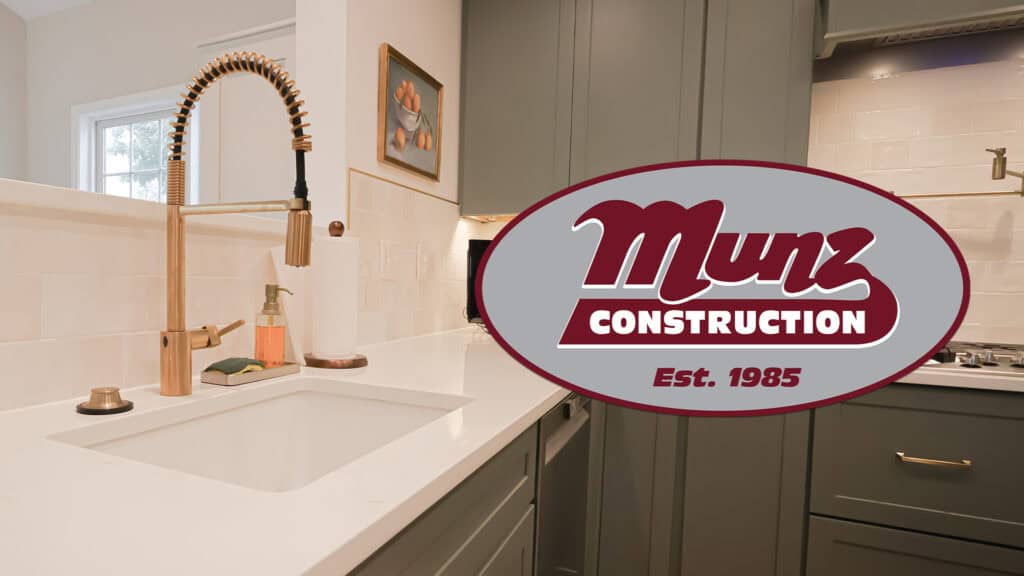 A modern kitchen with a gold faucet, white countertop, and gray cabinets, featuring the Munz Construction logo and Est. 1985 overlaid in the foreground—discover the best time to start a renovation with us.