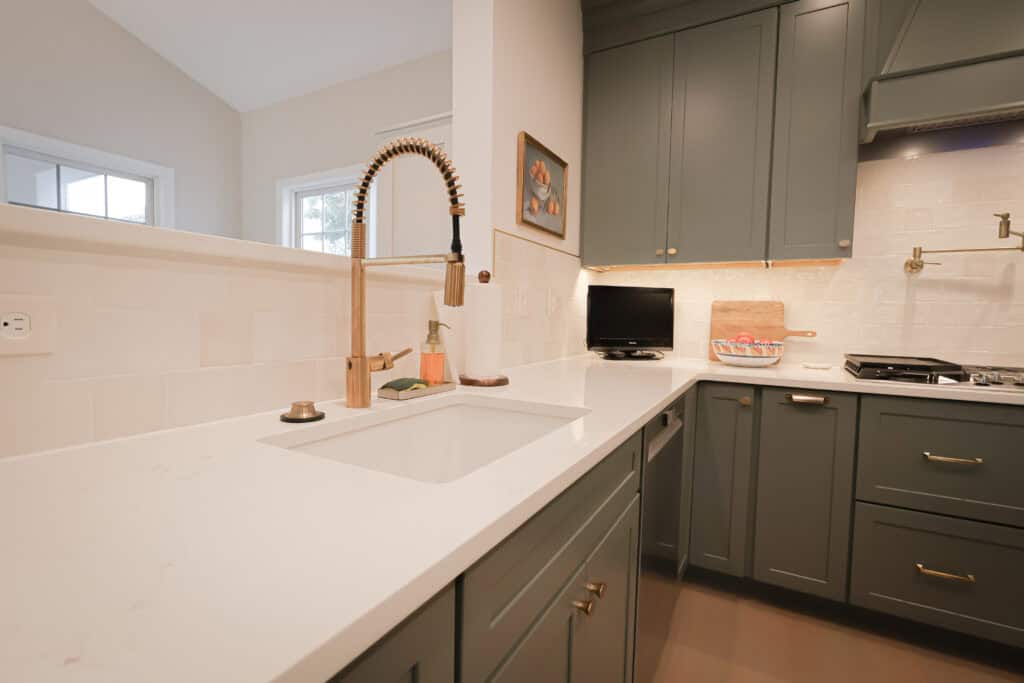 Modern kitchen with green cabinets, white countertops, a gold pull-down faucet, a small TV, a basket of fruit, and white tile backsplash. Natural light streams in—perfect inspiration if you're exploring how to add an in-law suite to your home.
