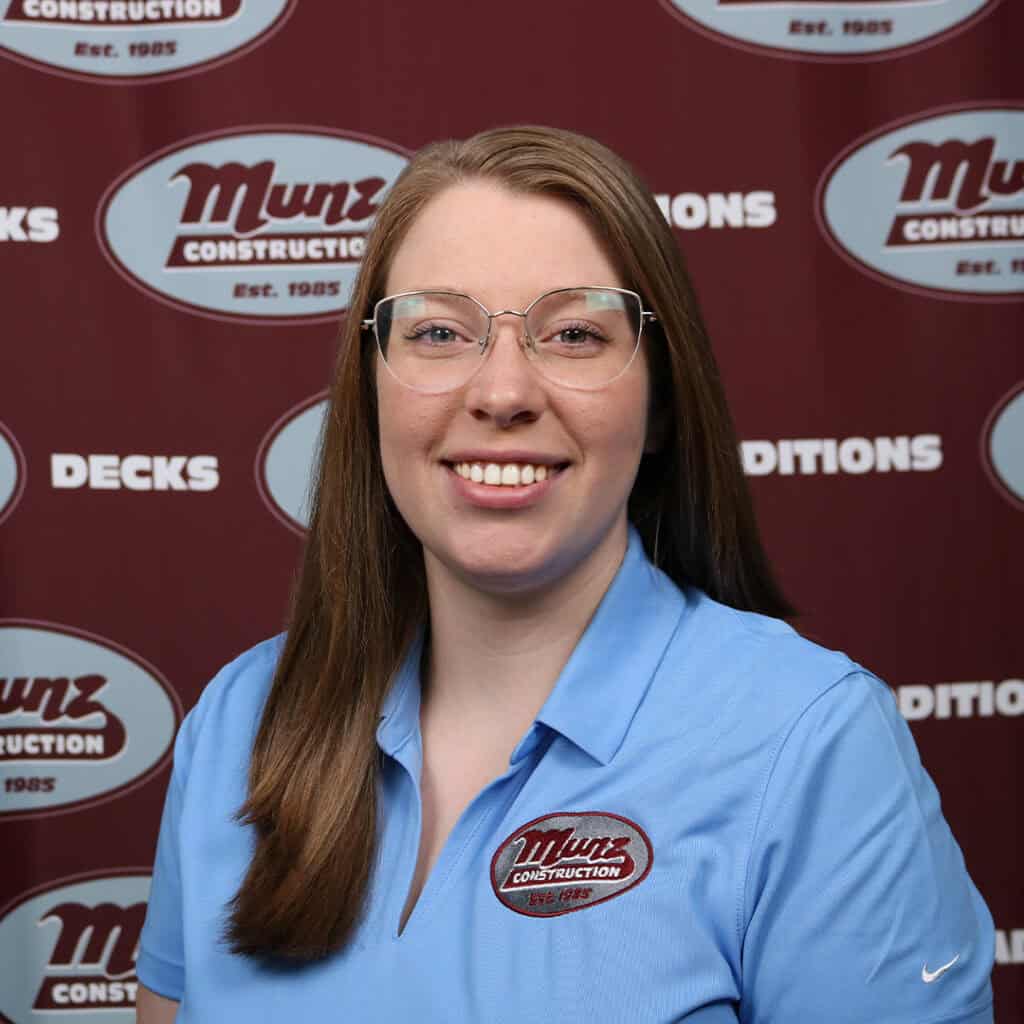 A woman with long light brown hair and glasses smiles at the camera. She is wearing a light blue polo shirt with a Munz Construction logo. The backdrop behind her displays the Munz Construction logo and words like DECKS and CONDITIONS.