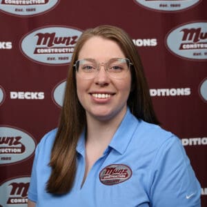 A woman with long light brown hair and glasses smiles at the camera. She is wearing a light blue polo shirt with a Munz Construction logo. The backdrop behind her displays the Munz Construction logo and words like DECKS and CONDITIONS.