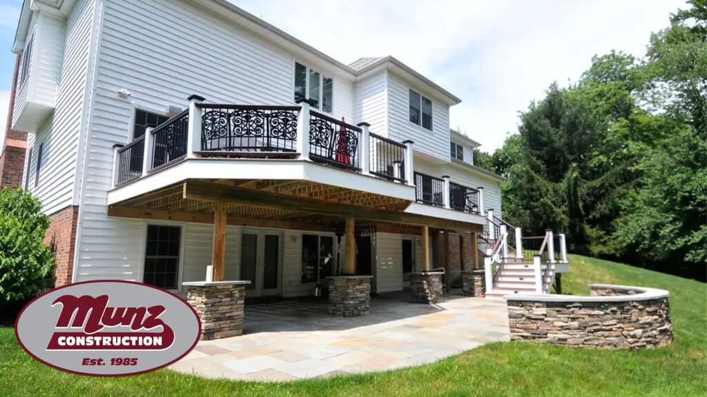 Back view of a two-story white house with a raised deck, stone pillars, and patio area. Thinking to plan a home addition in winter? Bucks County and Mercer County homeowners can find inspiration amid lush trees and grass. "Munz Construction Est. 1985” logo at lower left.