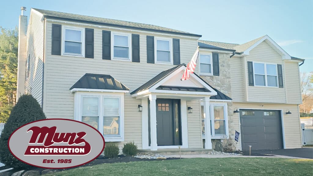A two-story beige house with black shutters, a stone accent wall, and a front porch. An American flag hangs above the entrance. The Munz Construction logo is in the lower left corner.