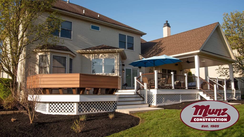 Two-level backyard deck attached to a beige house, featuring white railings, a blue umbrella, and landscaped garden beds—perfect for enjoying your Love Where You Live home improvement. The Munz Construction logo is in the bottom right corner.