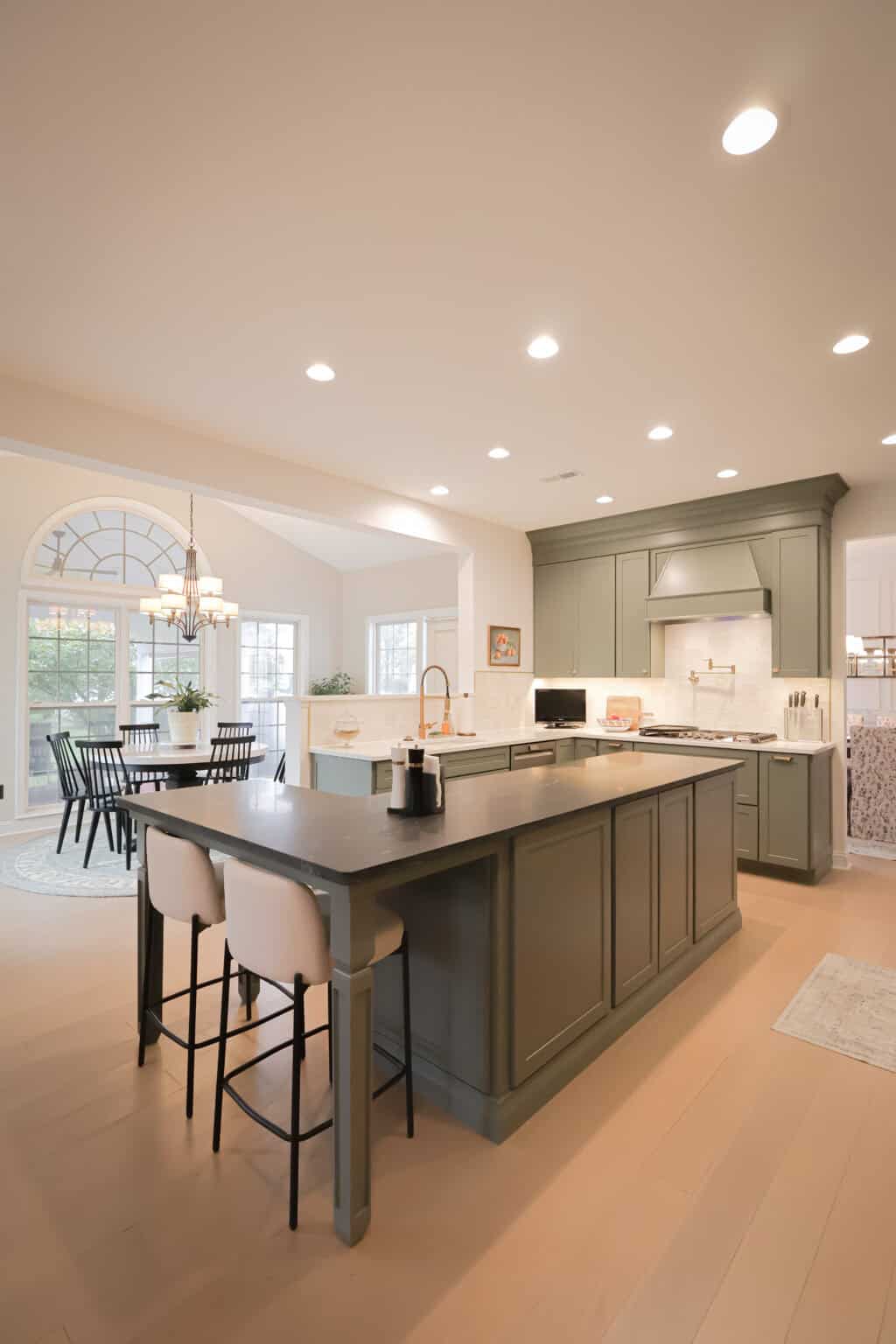 Spacious modern kitchen with sage green island, two barstools, built-in stove, and marble backsplash. Large windows and a dining table are visible in the bright, open dining area. Recessed lighting illuminates the room.