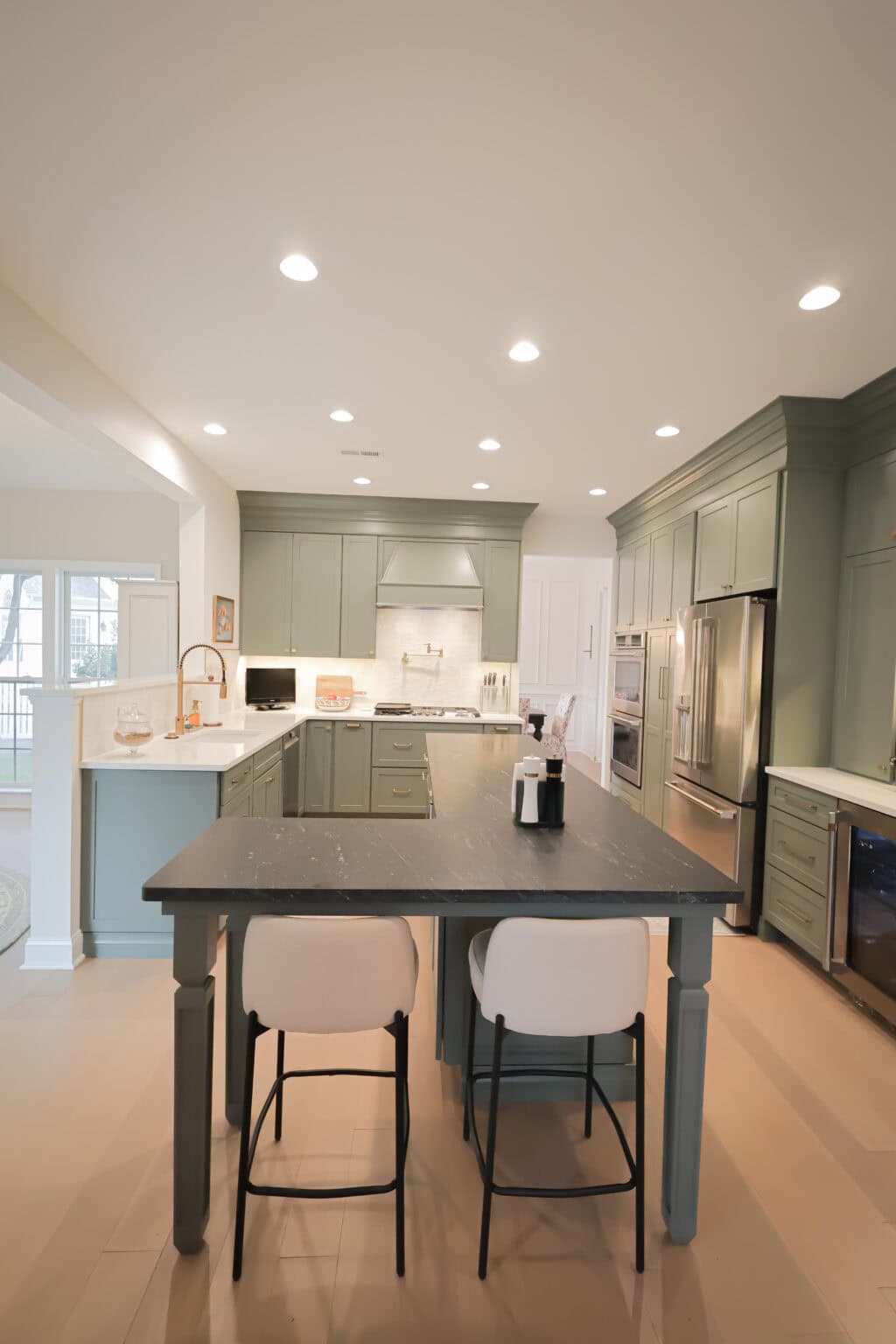 Modern kitchen with sage green cabinets, a large dark island with two white barstools, stainless steel appliances, under-cabinet lighting, and recessed ceiling lights. Light wood flooring and neutral walls complete the space.