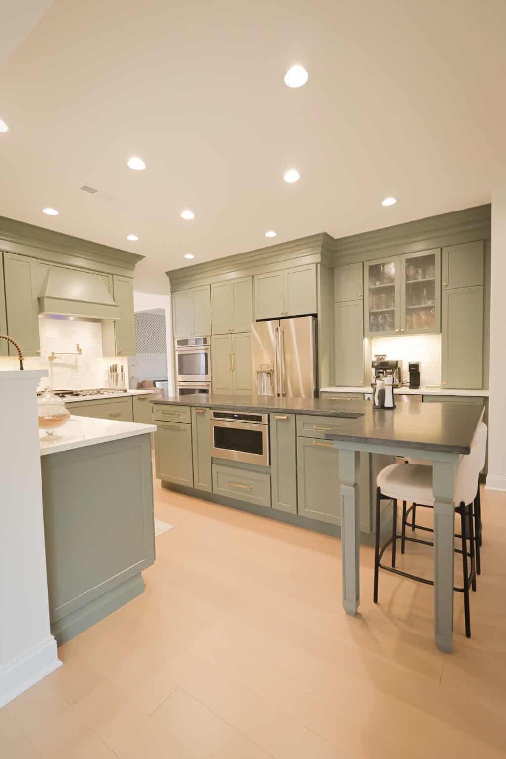 Modern kitchen with sage green cabinets, stainless steel appliances, a built-in oven, and a small bar-height table with two upholstered stools. Recessed ceiling lights brighten the light wood floor.