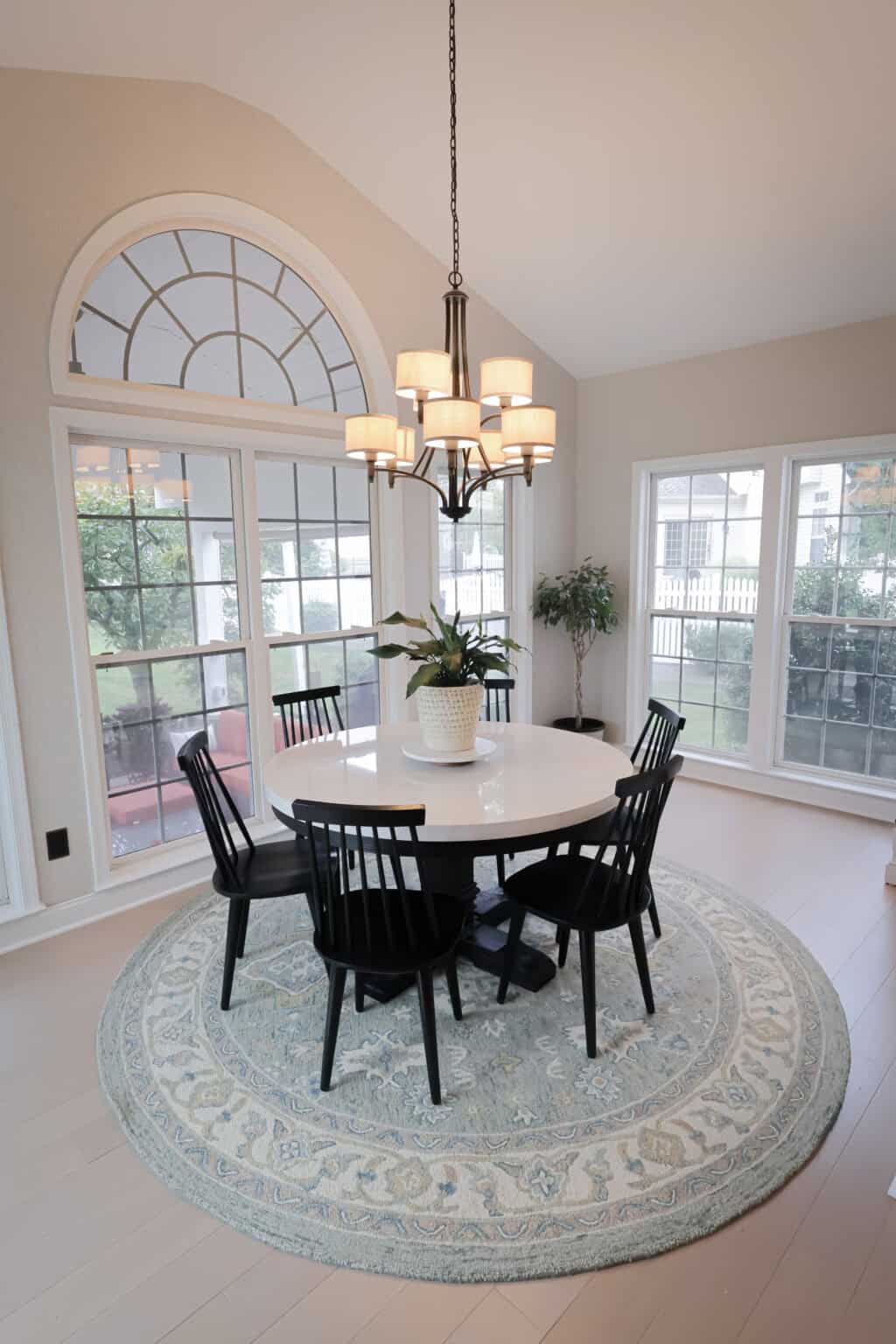 A dining room with a round white table, six black chairs, a potted plant centerpiece, a round patterned rug, large arched and rectangular windows, and a black chandelier hanging above.