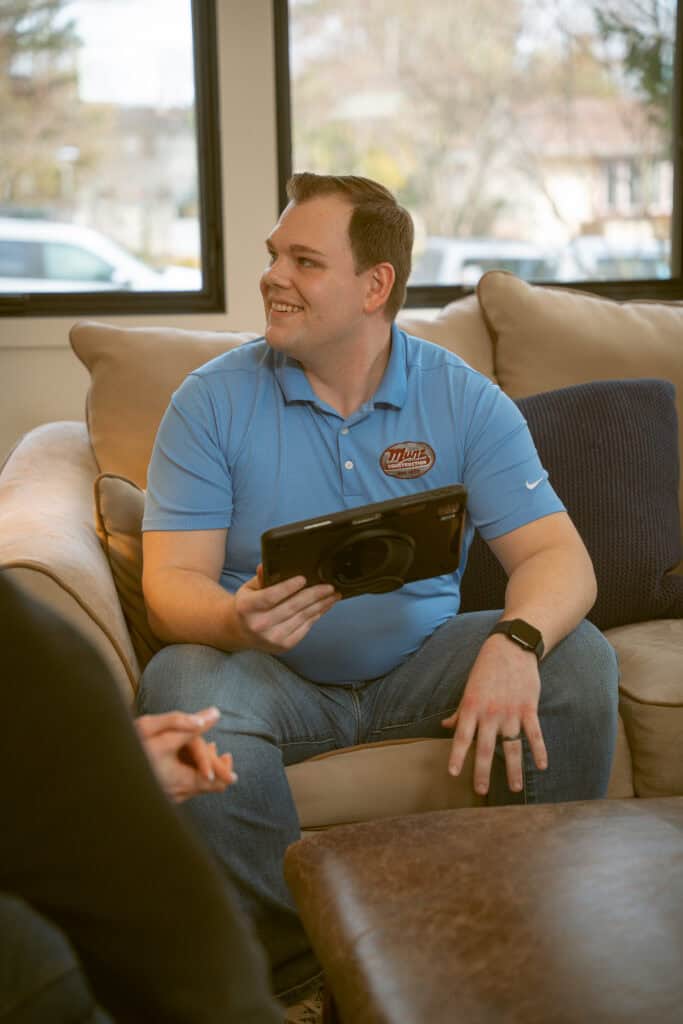 A man in a blue polo shirt sits on a beige couch, smiling and holding a tablet. Another person’s hand is visible in the foreground. The setting appears to be a casual living room with large windows in the background.