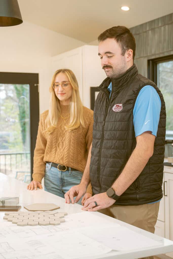 A woman and a man stand at a kitchen island covered with tile samples and design plans. The woman wears glasses and a brown sweater; the man wears a black vest and blue shirt. They appear to be discussing interior design ideas.
