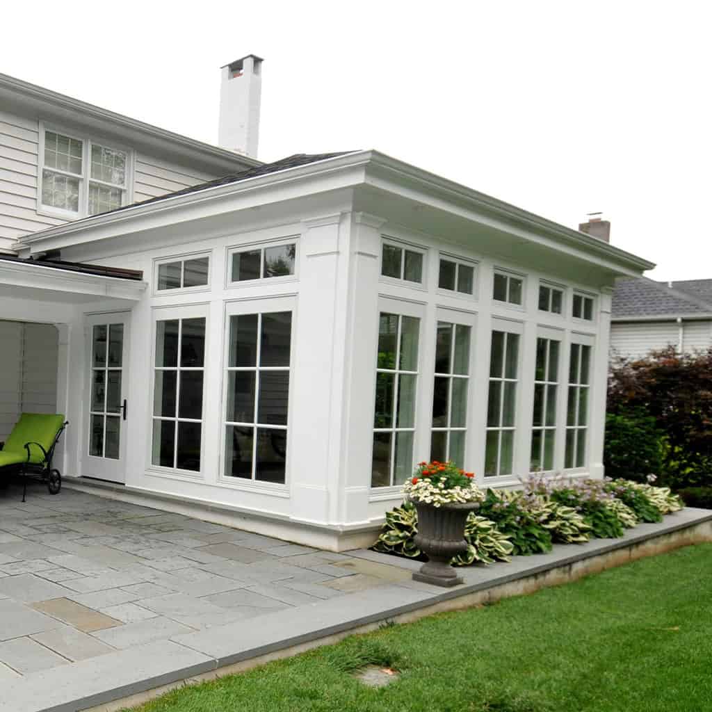 A white sunroom with large windows on all sides extends from a house, overlooking a stone patio with a green lawn and potted plants nearby.