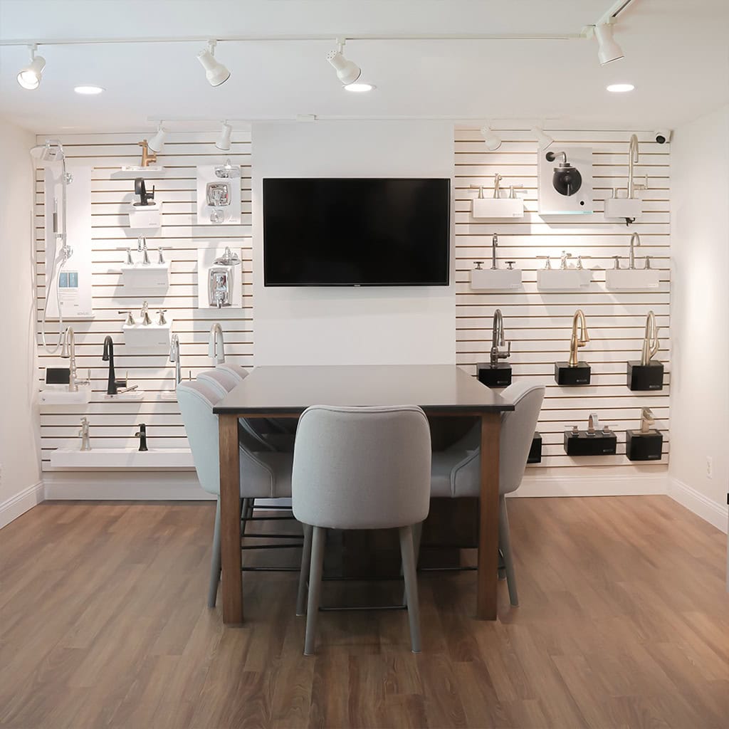 A modern showroom with a table and four chairs in the center, a wall-mounted TV, and displays of various kitchen and bathroom faucets on slatted white walls. The room has wooden flooring and track lighting.