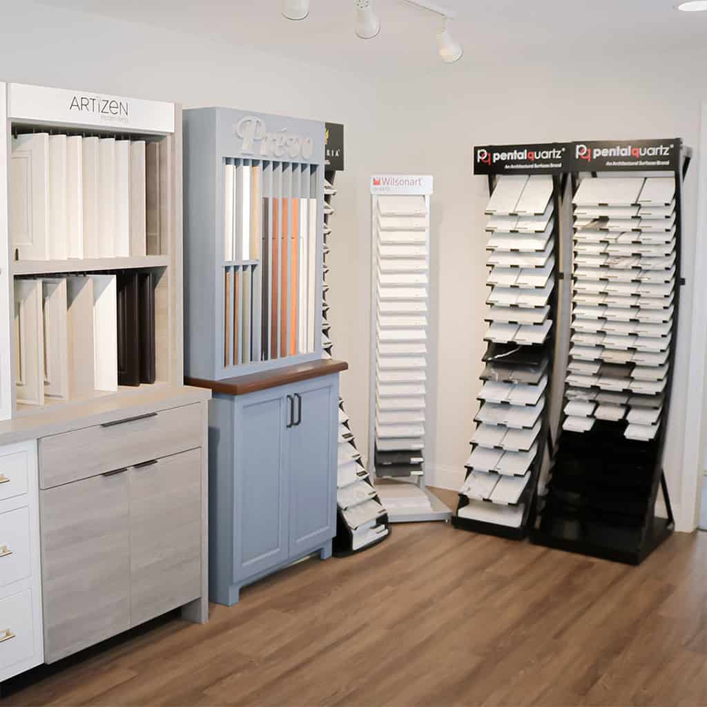 Showroom corner displaying sample boards of cabinetry, countertop, and quartz surface materials on stands, with a variety of colors and textures, set against a light-colored wall and wood flooring.