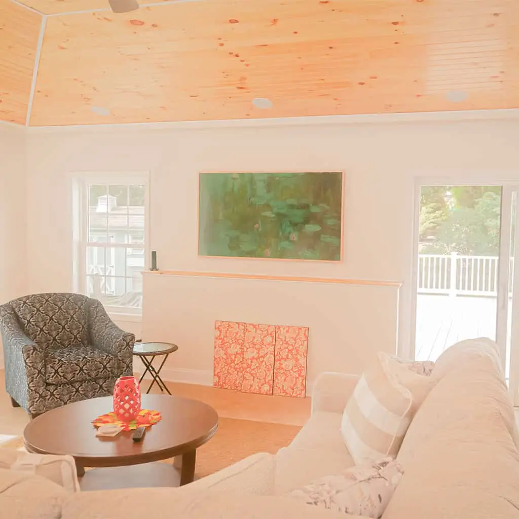 a bright living room with a white couch, patterned armchair, round coffee table, and large windows. a green painting hangs above two red patterned canvases on a shelf below a wooden ceiling.