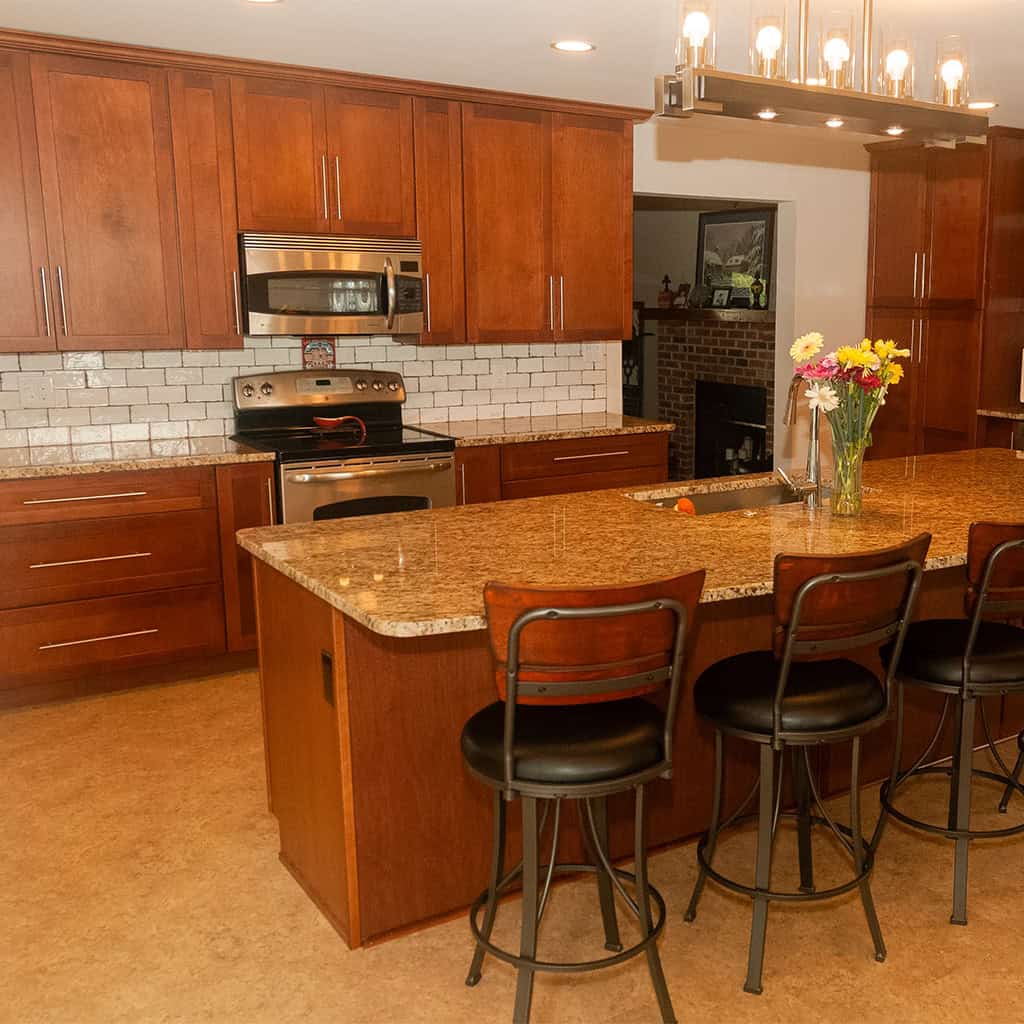 a modern kitchen with wooden cabinets, stainless steel appliances, a granite island countertop, three black and brown chairs, and a vase of flowers on the island. white subway tile backsplash and a light fixture overhead.