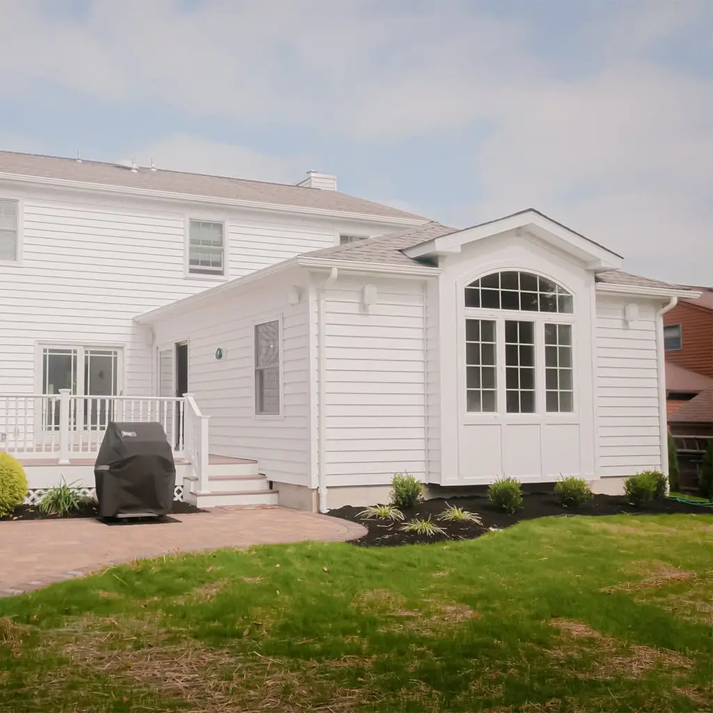 a white house with horizontal siding features a small back addition with large windows and a door, a paved patio area, and a grill cover beside the lawn and young landscaping under a partly cloudy sky.