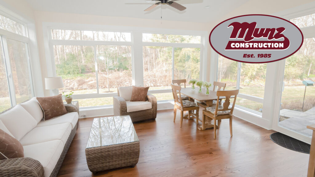 a bright sunroom with large windows, wicker furniture, a white sofa, armchair, wooden dining table with chairs, and hardwood floors—perfect for home upgrades for summer in bucks county. munz construction logo in the top right corner.