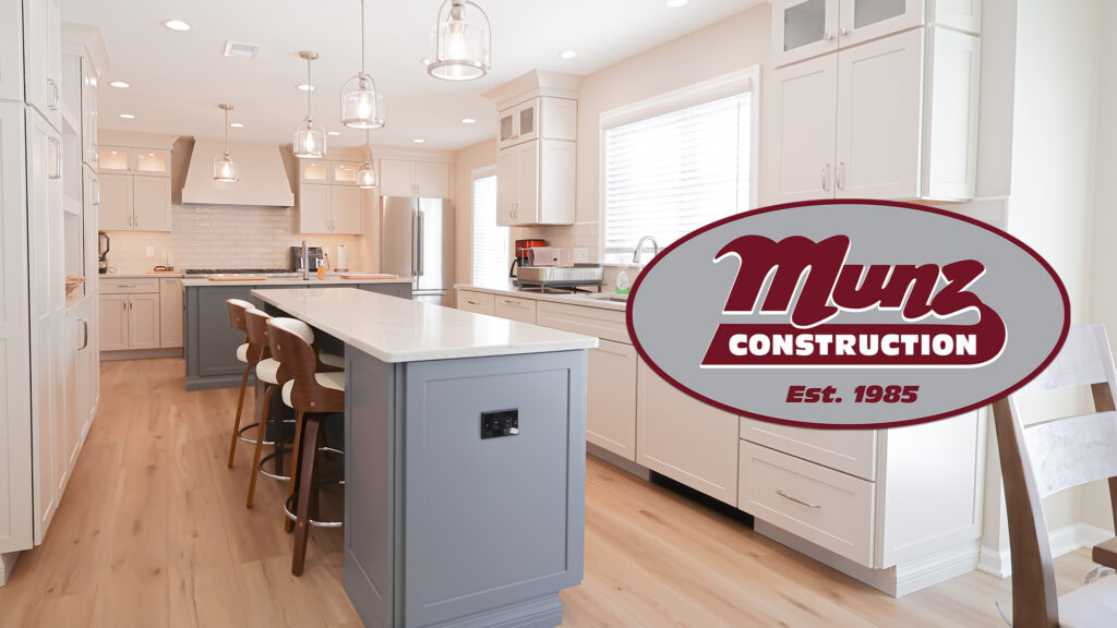 a modern kitchen with white cabinets, wood floors, a gray island with barstools, pendant lights, and munz construction’s maroon and white logo with est. 1985 overlaid on the right side.