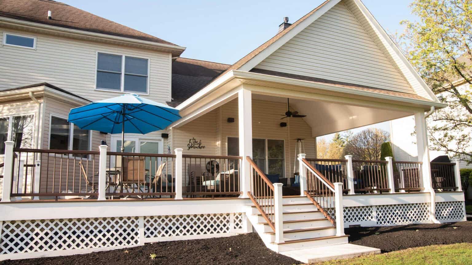 a large beige house with a covered back porch, white railings, and steps leading down to a mulched yard. patio furniture and a blue umbrella are on the deck under the afternoon sun.