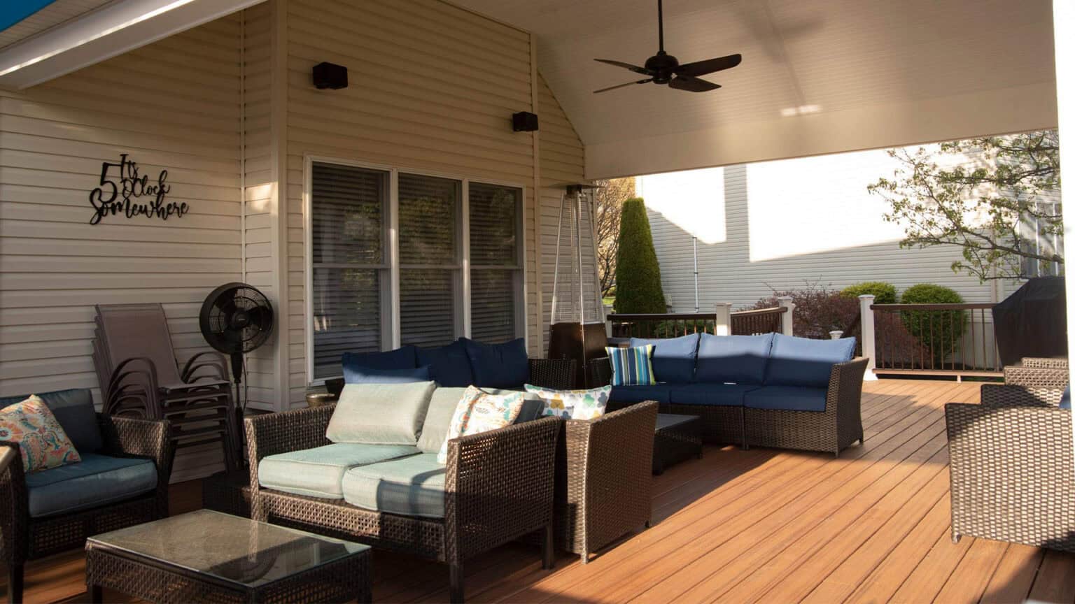 a covered outdoor patio featuring wicker furniture with blue cushions, decorative pillows, a glass top coffee table, a standing fan, and a wall sign reading the smith sunhouse. sunlight filters onto the wooden deck.