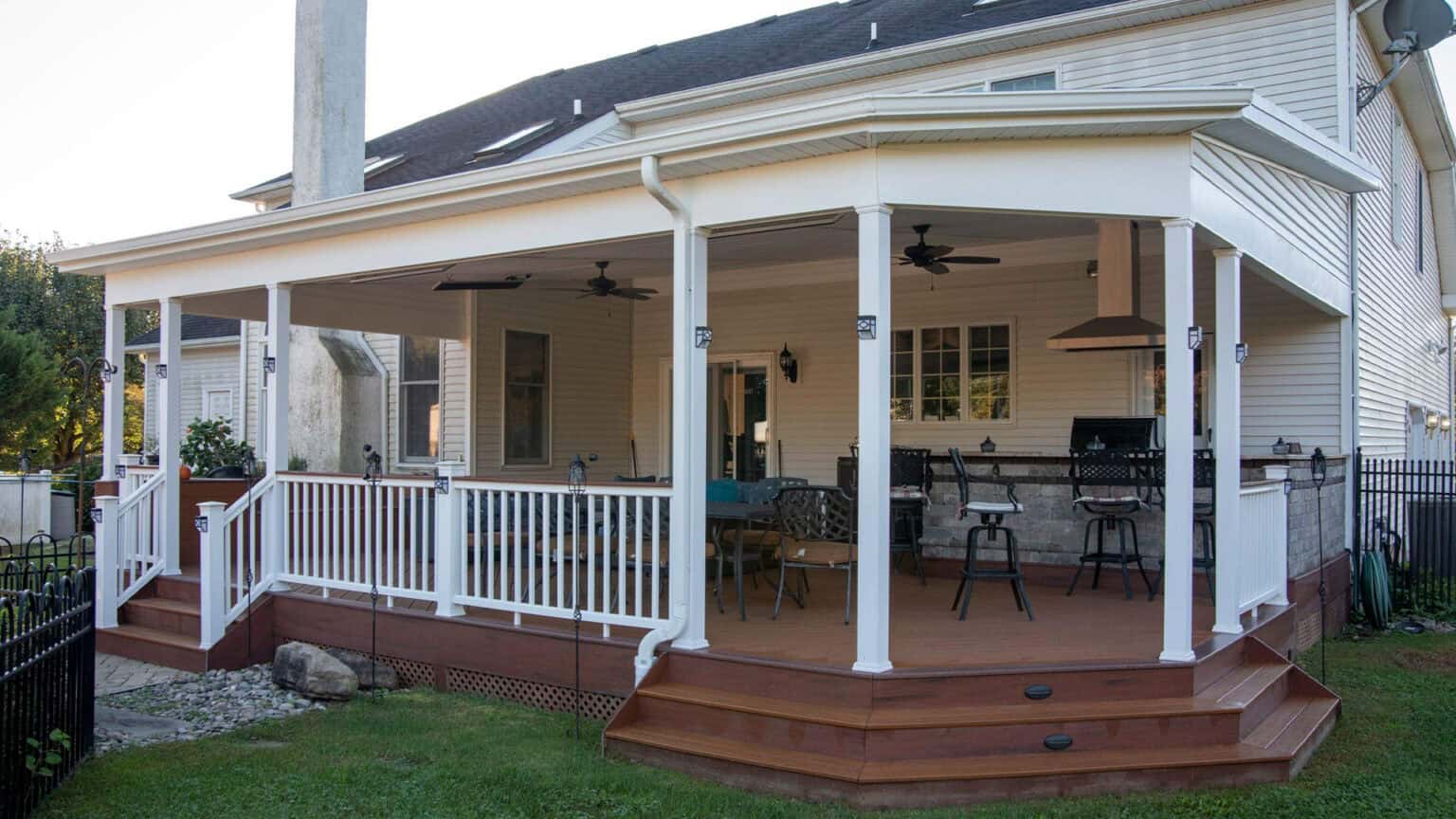 a spacious covered backyard porch attached to a beige house, featuring white railings, brown wooden floor, outdoor furniture, ceiling fans, and an outdoor kitchen area with barstools. green lawn surrounds the porch.