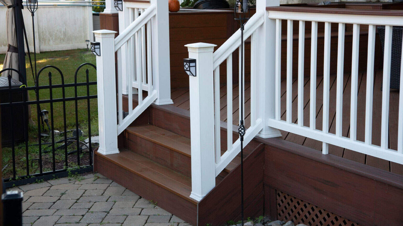 a small porch with brown wooden steps and white railings leads up from a paved patio area to a deck. decorative black lanterns are mounted on the railing posts. grass and a black fence are visible in the background.