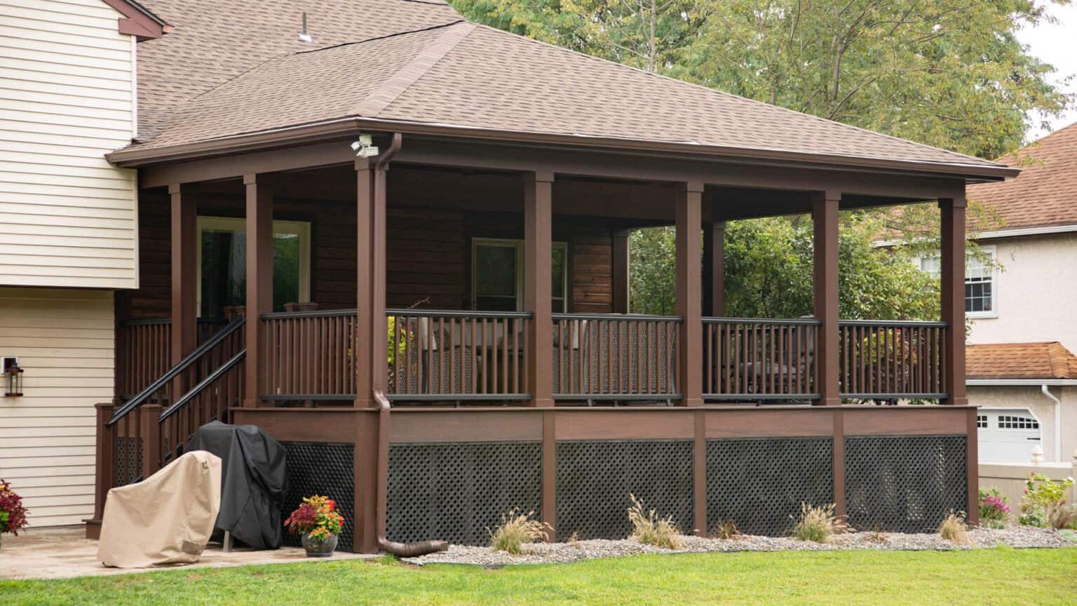 a screened in, elevated porch with wooden railings and stairs attached to a house, featuring covered grills and potted plants nearby, set in a green backyard.