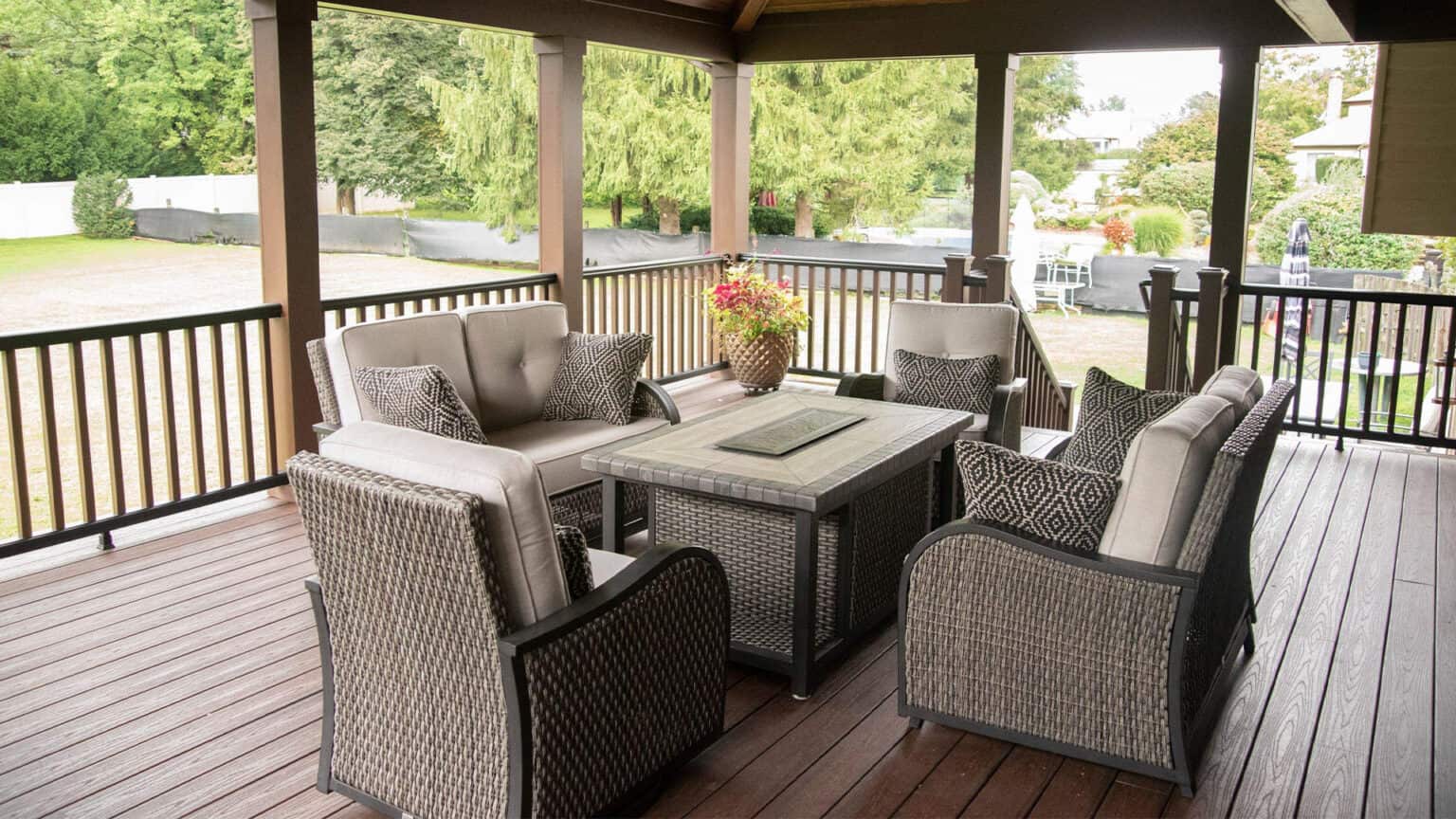 a covered outdoor patio with wicker furniture, including cushioned chairs and a central table with a flower pot. the patio overlooks a spacious yard with trees and a white fence.